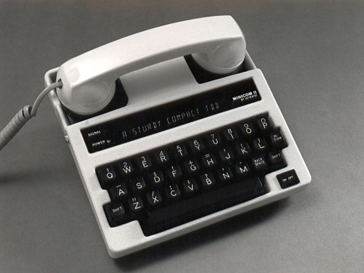 Ultratec Minicom II TTY device: a light-gray text telephone with a handset cradled on top and a black QWERTY keyboard below.