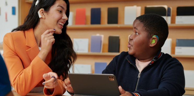 A woman wearing a hearing aid showing a boy wearing a hearing aid how to use an iPad.